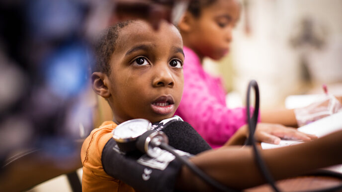 A young boy with a blood pressure cuff around his arm looks up at a health professional reading the dial