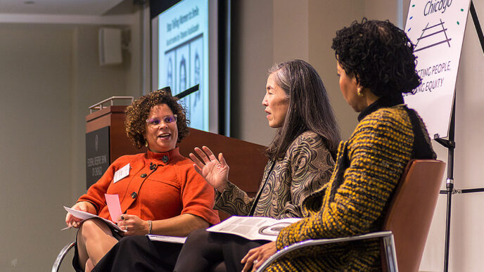 Three women sit in chairs in a stage.