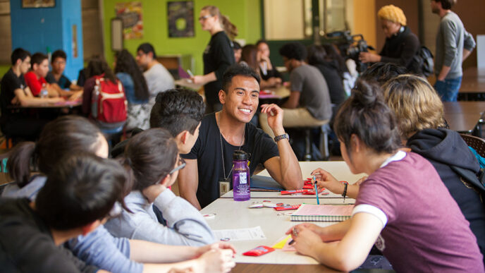 A smiling young man seated around a table with a group of high school students.