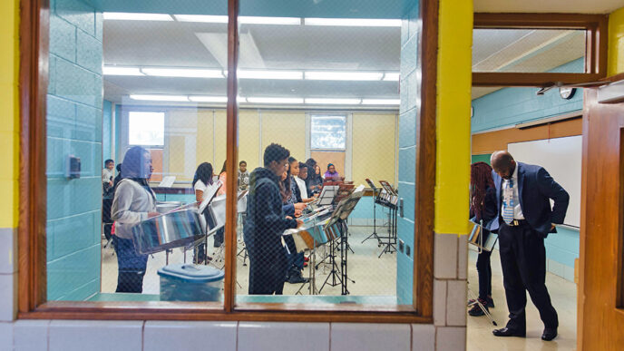 Students rehearse in a music classroom at Whistler Elementary School|A young musician holds a mallet poised over a steel drum|Julian Champion conducts a music class|Two girls in the percussion section read sheet music together