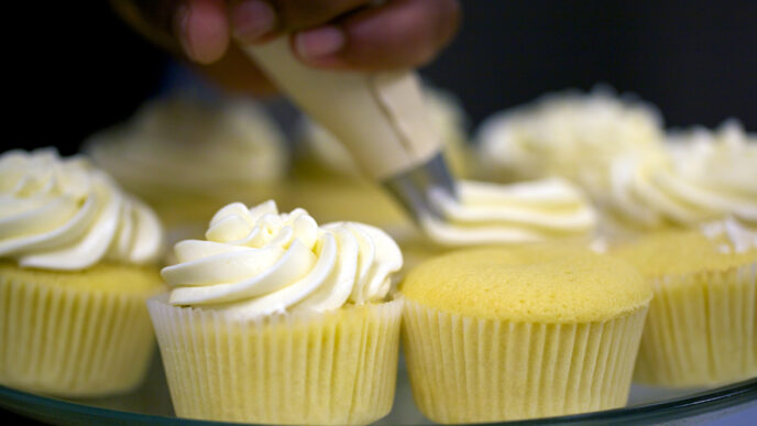 A baker pipes swirls of vanilla frosting onto a platter of cupcakes|Lisa Thompson serving up a cupcake and smiling|Lisa Thompson working alongside a trainee in the bakery's kitchen|