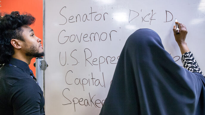 Mahjeed Marrow with a student from his citizenship class write on the classroom whiteboard|Mahjeed Marrow waits for a train on the Red Line station platform|A young boy plays a computer game in the office at the Pan-African Association|A student fills out a practice test of citizenship questions|Mahjeed Marrow helps a student complete a list of elected officials