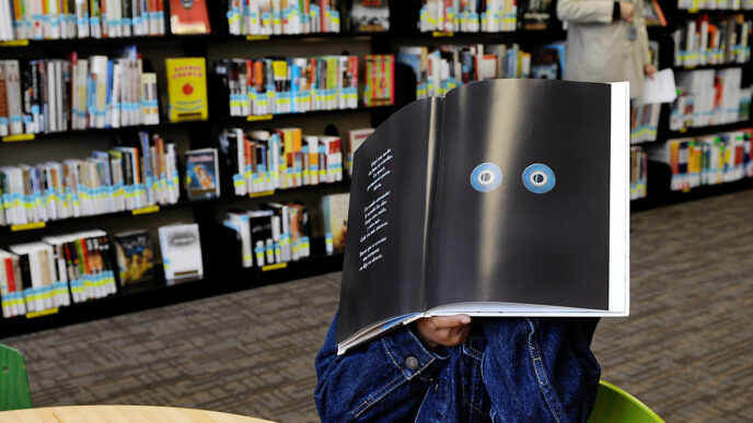 A boy sitting in a library holds a picture book in front of his face|Deborah Summers