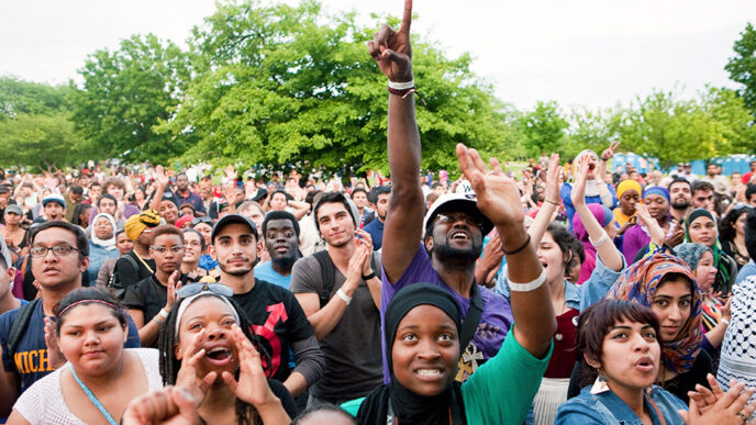 Rows of people seated in folding chairs and standing at an outdoor concert clap and cheer