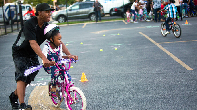 A young girl pedals a bike while a man holds the handlebars and helps her steer