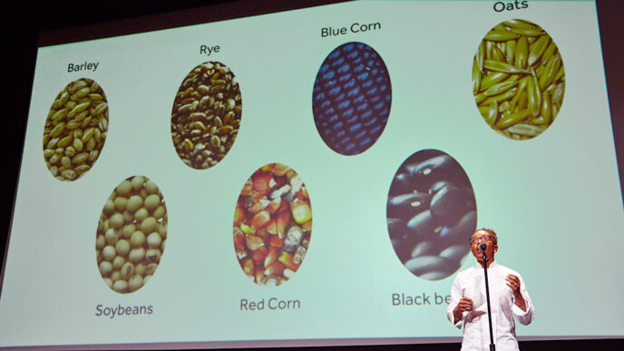 A chef gives a stage presentation about beans and grains|A display of garlic and root vegetables on a table|Four people on stage in front of a projection screen|Members of Team Leverage hugging and smiling|A group of judges seated at a table while one speaks into a microphone|Crowded auditorium at the Museum of Contemporary Art|