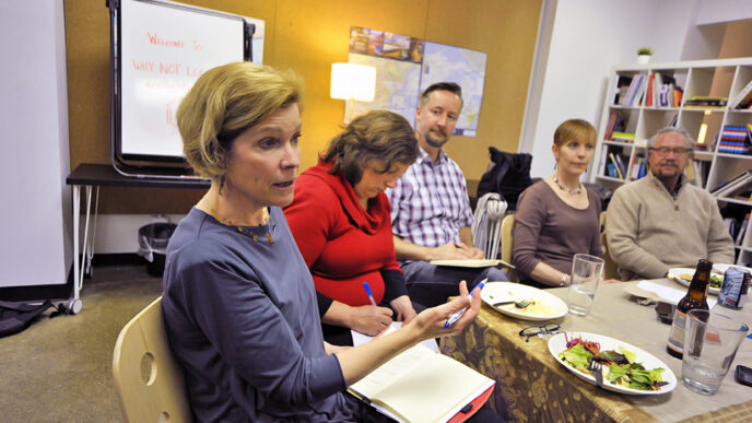 Group of people seated around a table talking|Whiteboard with the words Welcome to Why Not Local?|Closeup of metal picture frame and desk accessories|Susan Page Estes leaning forward listening to conversation|View from above of thirteen people seated around a table talking