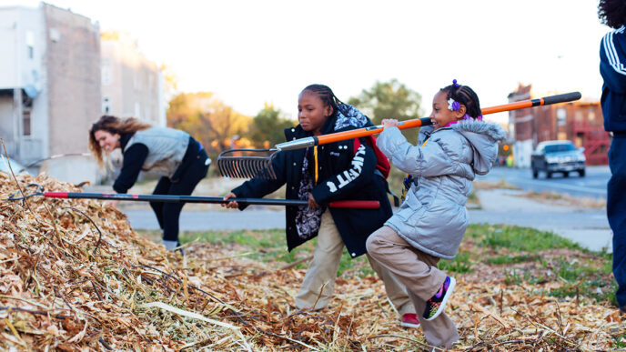 Two young girls raking wood chips in a garden|A colorfully painted stump with handprints and the words Black Lives Matter|A garden educator listens to a conversation|A young woman leans on a rake