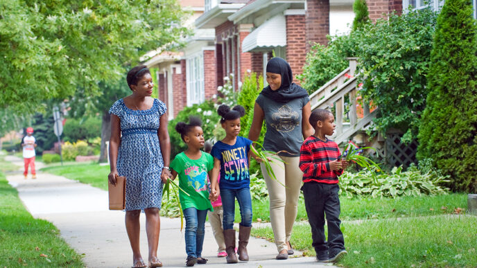 Two women and three young children walk along a sidewalk in a residential neighborhood|Two women seated at a table in conversation|A woman holds a toddler on her lap as they look out a window