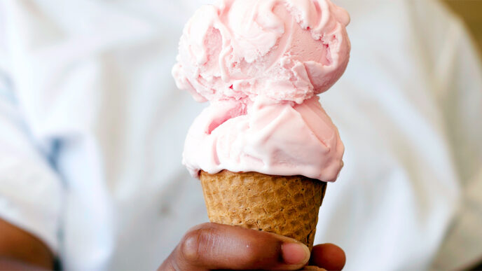 An ice cream server holding a cone with two scoops|Hand scooping ice cream from a freezer case|Jeni's Storefront on Southport Avenue in Chicago|Three people sitting at a table holding waffle cones
