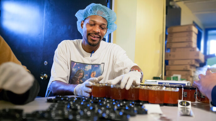 A man seated at a workbench seals jars of honey