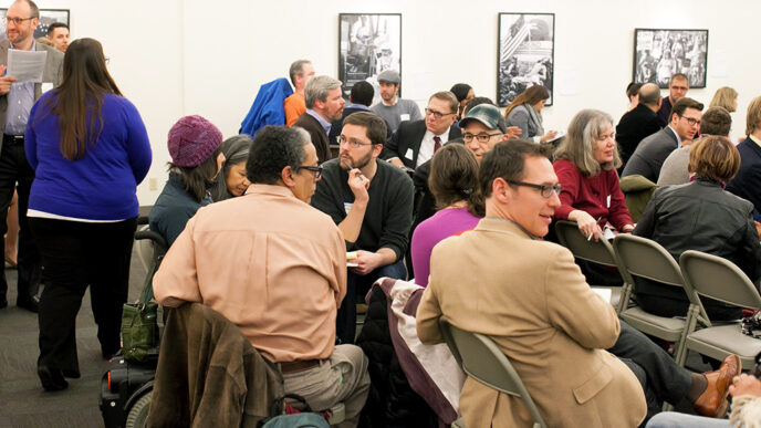 A group of people seated in chairs and motorized wheelchairs in an animated discussion|A speaker seated behind a table addresses a meeting using a microphone