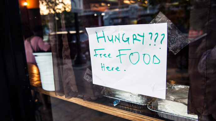 A hand-written sign taped on a restaurant window reads Hungry? Free Food Here|A A man seated at a table speaks and gestures to several women seated beside him||A woman tapes a sign onto the window of a restaurant|A smiling woman with her arms folded|Four people fill their plates from chafing dishes on a buffet table|A man gestures and speaks intensely while people in the background listen|A young woman speaks to a man seated at a table beside her