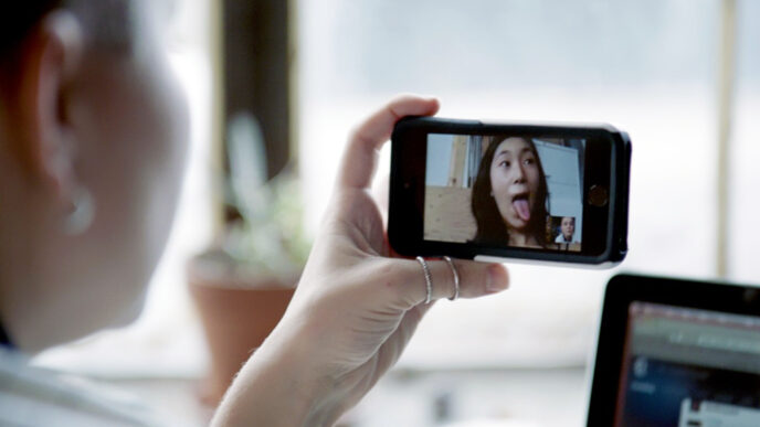 A young woman's hand holding a phone to record herself on video; the screen shows her sticking her tongue out in a playful face