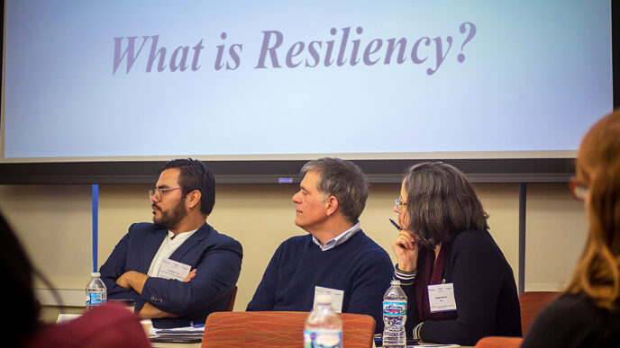 Three people steed below a projection screen displaying the words What Is Resiliency?|Several dozen people in a conference room face a row of panelists in conversation