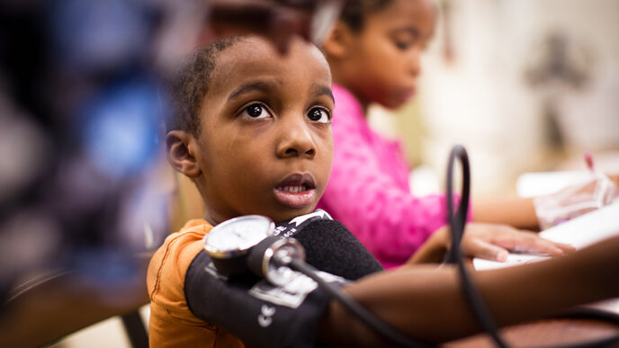A young boy with a blood pressure cuff around his arm looks up at a health professional reading the dial