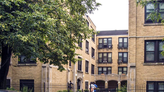 A brick apartment building with a central courtyard and large trees in front|Two young children sit on beds with brightly colored bedspreads
