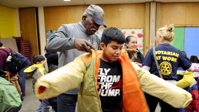 A man wearing a baseball cap helps a boy put on a new tan winter coat|A small girl sits at a table beside her mother