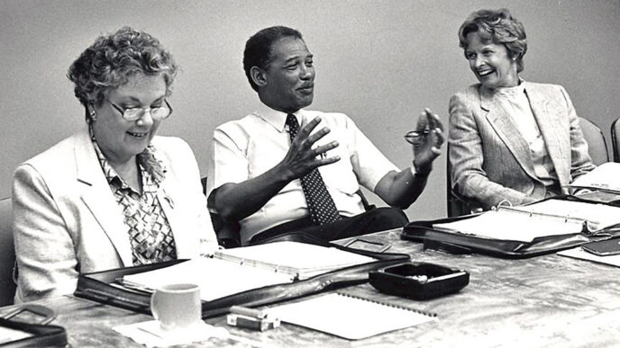 Black & white archival photo of a meeting at the Trust's offices. At a conference table covered with binders and papers