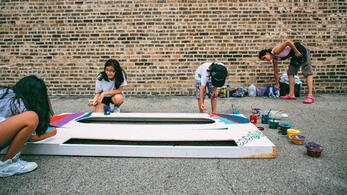 Three teenagers paint a large mural panel lying on the ground