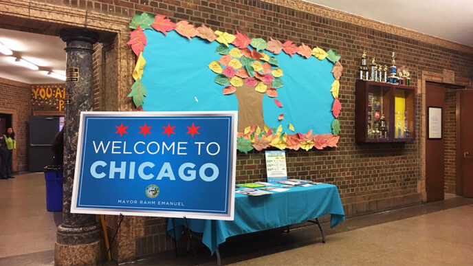 An easel in a large gymnasium holds a large sign reading Welcome to Chicago|A woman seated at a table speaks and gestures with her hands|Five people seated around a table in front of a projection screen