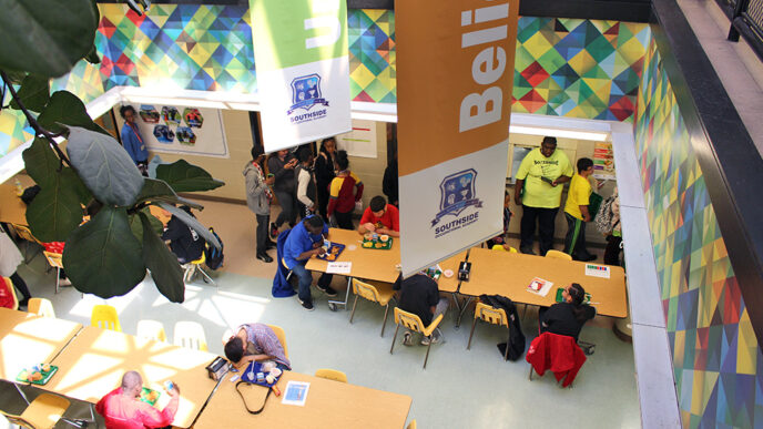 Aerial shot of the Southside Occupational Academy cafeteria. Students gather to have lunch and converse at the cafeteria tables.|A worker wearing a chef's jacket and sequined cap pushes a tray of ingredients through a commercial kitchen|Five people smile to the camera in front of a sign reading Southside Occupational Academy - Welcome