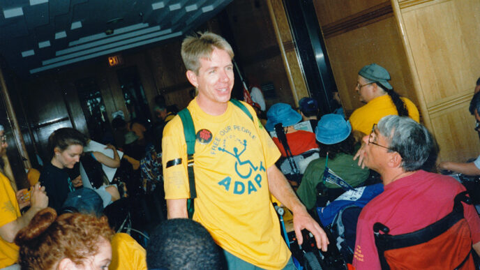 A man wearing a t-shirt that says ADAPT amid a group of people using wheelchairs|Vintage photo of protestors shouting and holding signs