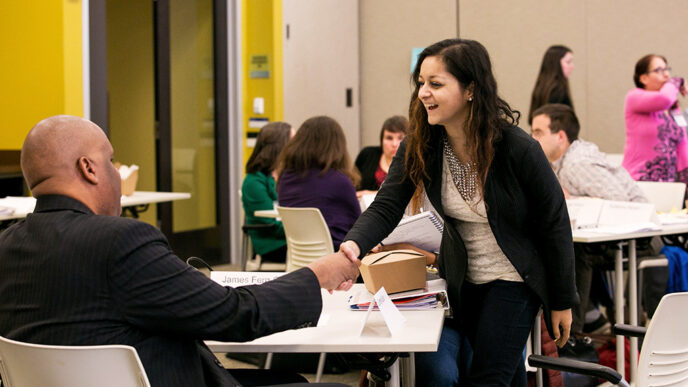 A standing woman smile sand shakes the hand of a man seated in a chair|A meeting room with people seated around shared tables