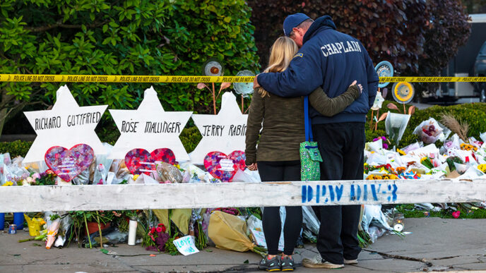 A man wearing a jacket that reads Chaplain embraces a woman as they stand in front of a row of memorials on a sidewalk|I Gave to #LoveIsStronger|The Pittsburgh Steelers team logo reworked to include a Star of David
