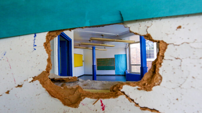 A hole in an interior wall shows a view into an empty room beyond|A woman stands with arms crossed