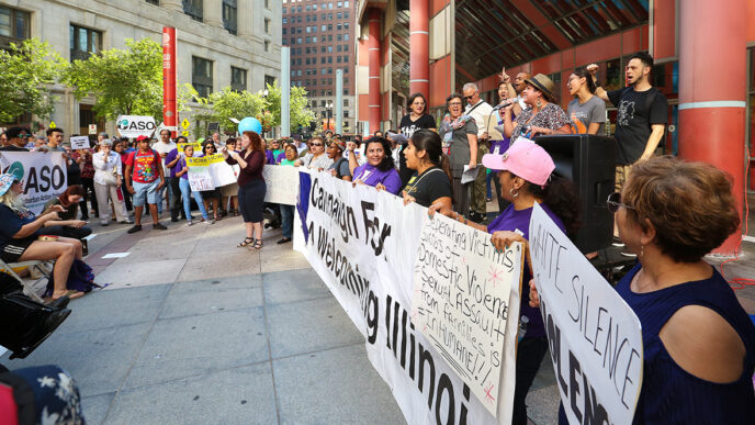 Speakers hold signs at a rally on the plaza surrounding a municipal building|A rally attendee holds up a sign reading I Am My Sister's Keeper|A group of women hold colorful hand-lettered signs in English and Spanish in support of DACA|Viewed from behind