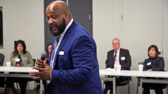 A man wearing a blue suit stands in front of a town of people seated at a conference room table