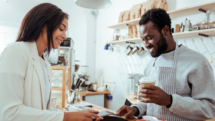 A smiling woman uses her phone to make a purchase at a coffee shop from an employee wearing an apron