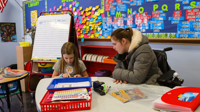 A teacher watches a student reading at a table in a brightly decorated classroom|A group of young children sit cross-legged on the floor facing a person using a wheelchair