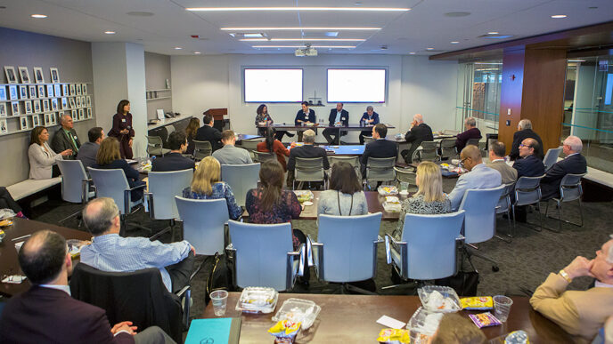 Four people seated at the front of a conference room face an audience in rows of chairs|A row of seated people