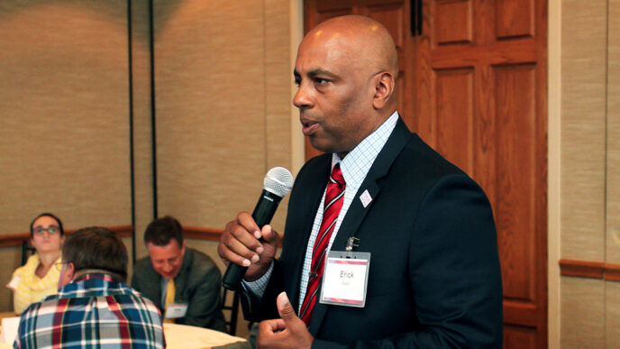A man holding a microphone speaks to people seated in a conference room