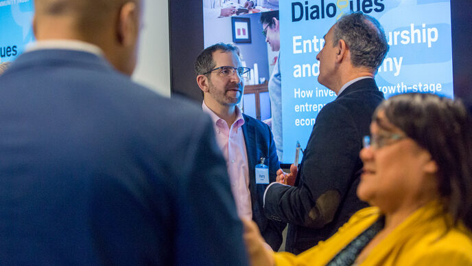Two pairs of people in conversation standing in front of a projection screen|A row of people seated at a conference table lean to see and listen to a speaker|Around a conference table