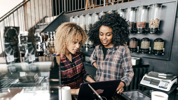 Two women behind the counter at a coffee shop look at a laptop